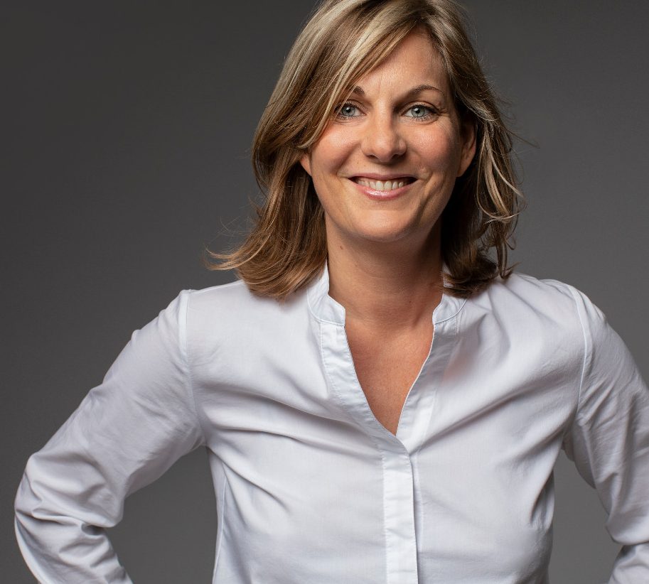 Foto Katrin Bohlen Lächelnde Frau mit schulterlangen Haaren, im schwarzen Blazer, die Arme verschränkt.